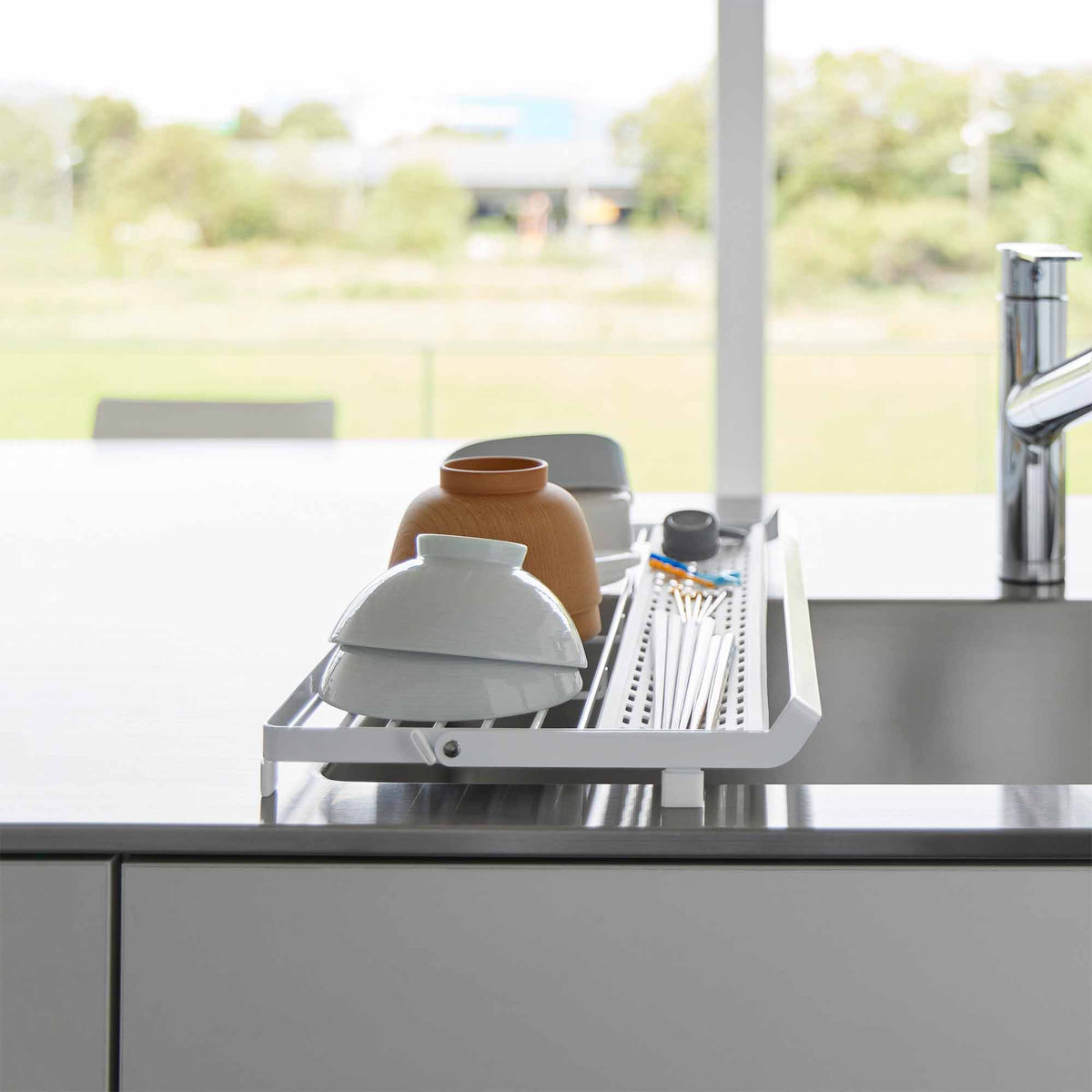 End view of the white Yamazaki Home Slim Over-the-Sink Dish Drying Rack - Steel spanning a sink, with bowls and cups arranged beside the perforated utensil section near a bright window.