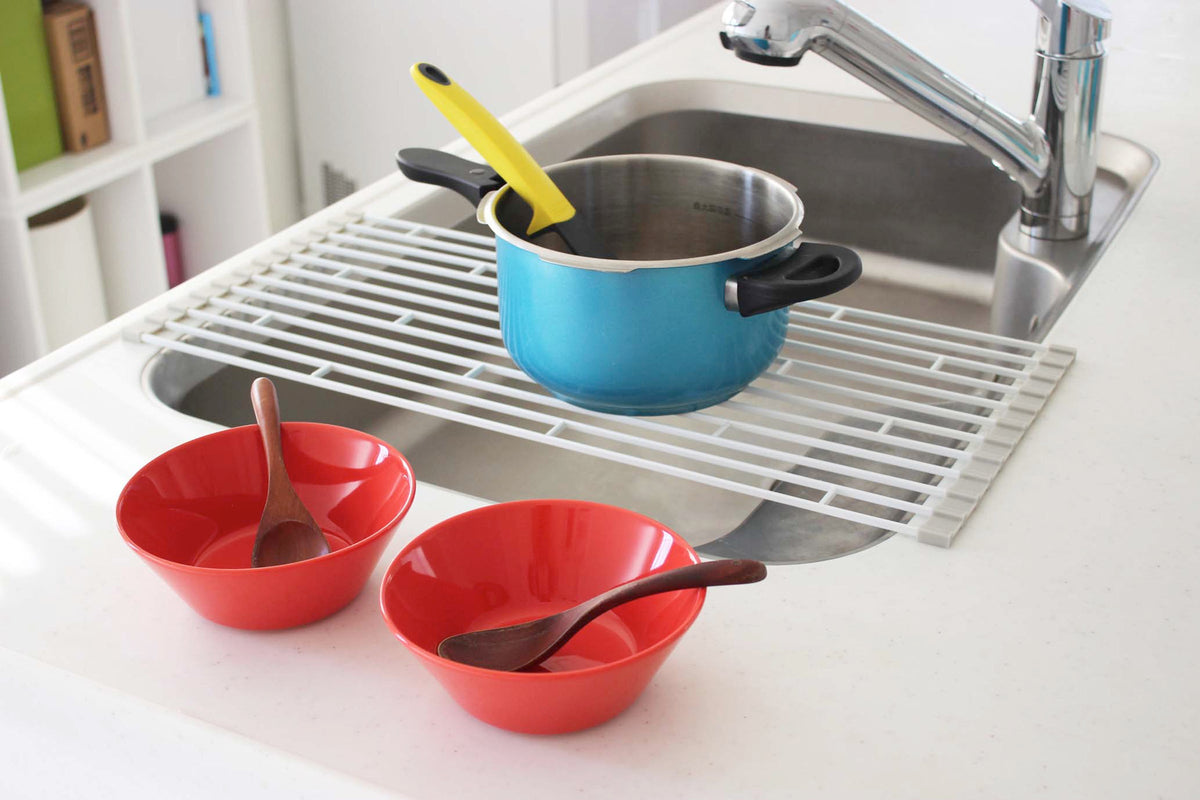 Yamazaki Home Over-the-Sink Dish Drainer - Steel spread across a sink beneath a blue saucepan with a yellow-handled utensil, with two red bowls and spoons on the counter in front.