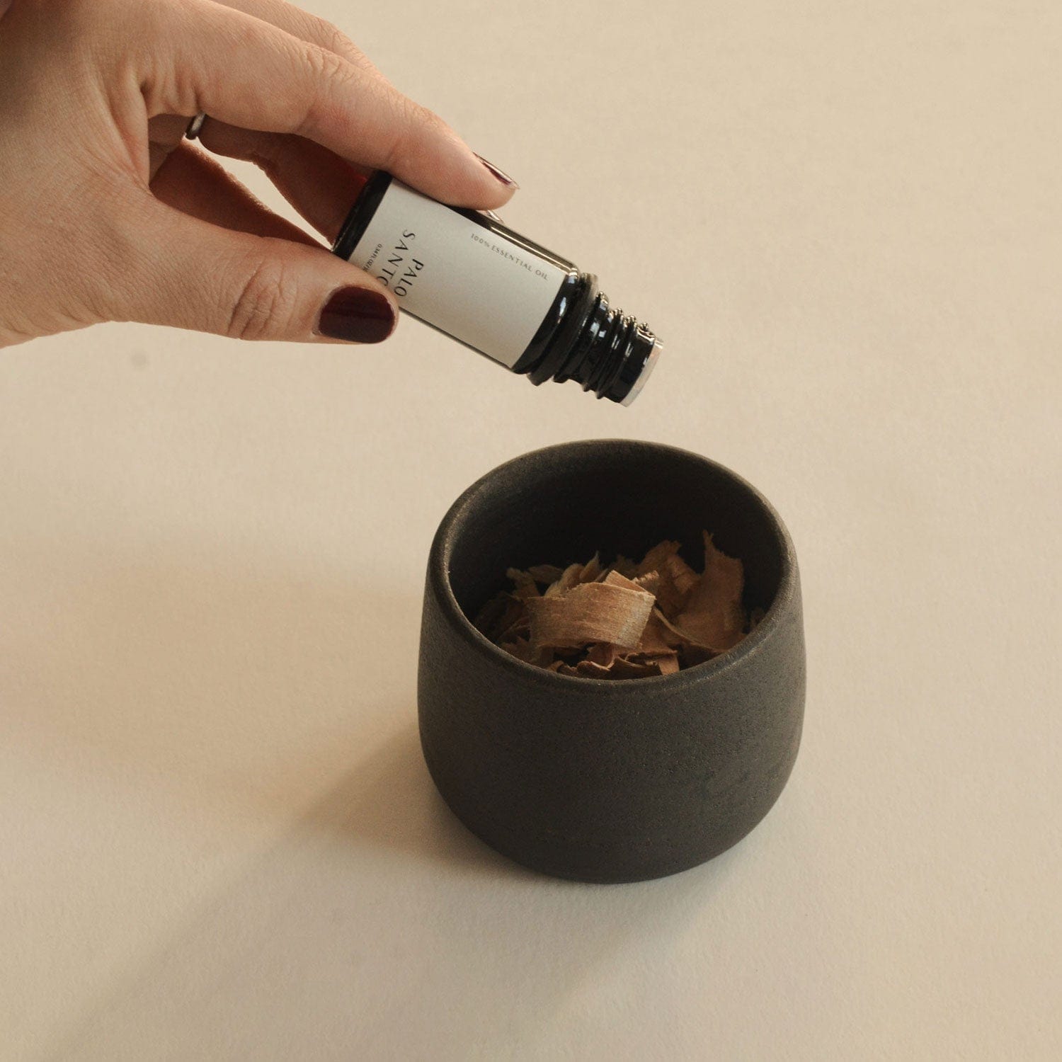 Hand holding a small bottle over a black cup filled with dried herbs on a beige background