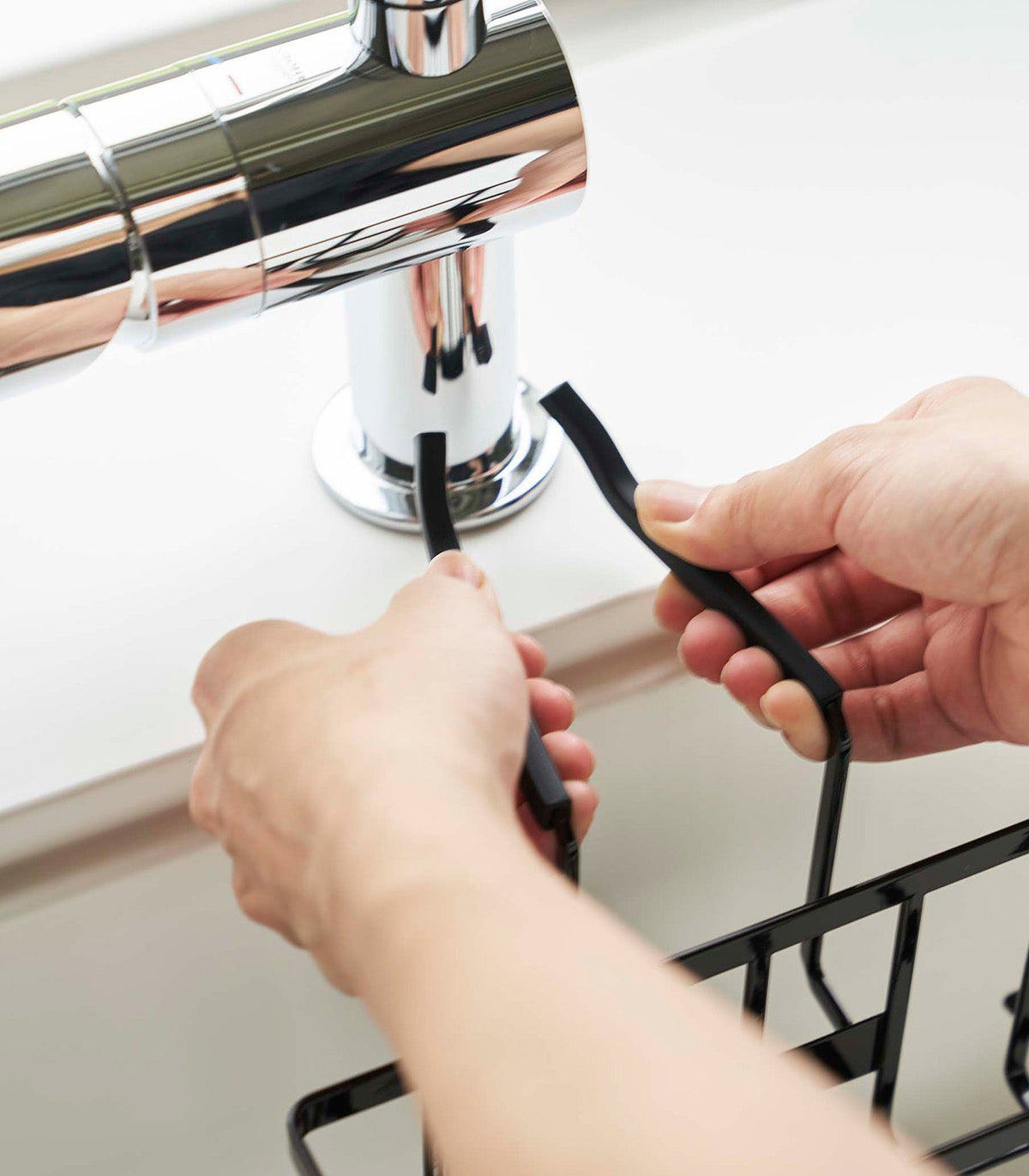 Two hands fit the black support arms of the Yamazaki Home Faucet-Hanging Sponge Caddy - Steel around the faucet base above the sink.