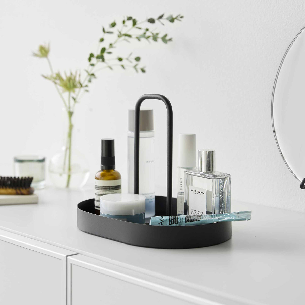 Black Yamazaki Home Vanity Tray with Handle - Steel organizing skincare containers, a clear perfume bottle and a toothbrush on a white bathroom counter beside a mirror.