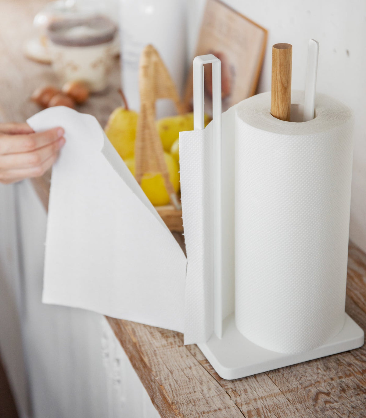 Wider kitchen view of a person drawing a sheet from the Yamazaki Home Paper Towel Holder - Steel + Wood, with its white rectangular base, tall side rail and wooden post visible beside lemons and cutting boards.