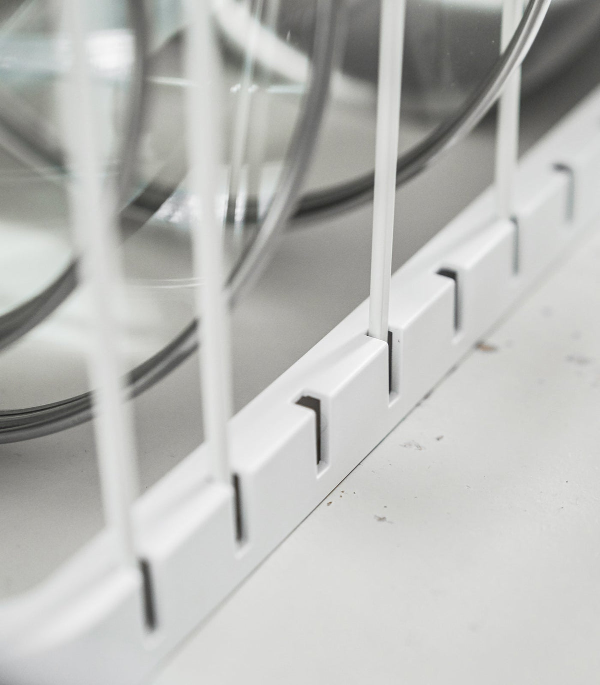 Close-up of the white Yamazaki Home Adjustable Pot Lid Organizer - Steel base, showing divider wires inserted into evenly spaced rectangular slots beneath upright lids.