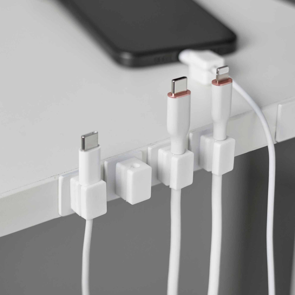 White Yamazaki Home Magnetic Cable Holders With Adhesive Steel Bases (Set of 5) - Silicone along a desk edge, holding charging connectors upright beside a black phone, with white cables hanging below.