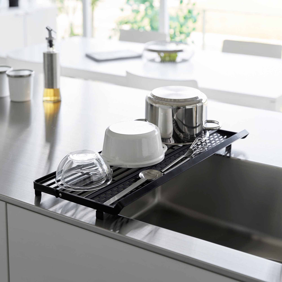 Black Yamazaki Home Slim Over-the-Sink Dish Drying Rack - Steel in a flat configuration over a sink, supporting an inverted saucepan, white bowl, clear glass bowl and cutlery.