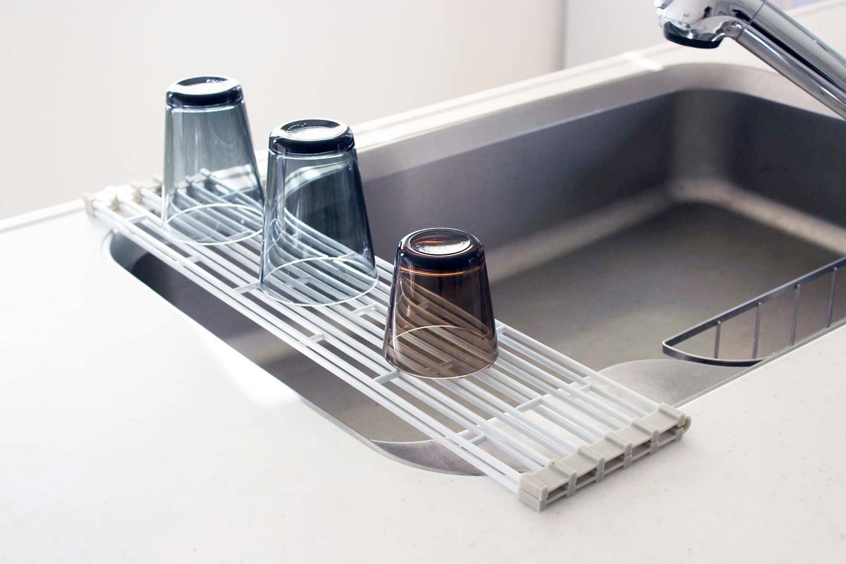 White Yamazaki Home Over-the-Sink Dish Drainer - Steel partially covering a stainless-steel sink, with three inverted blue and brown drinking glasses resting on its parallel bars.