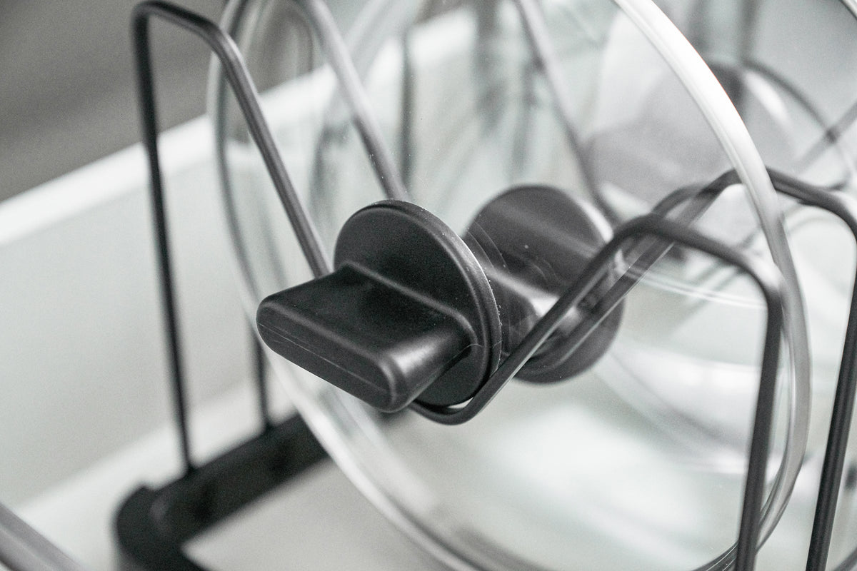 Close-up of the black Yamazaki Home Adjustable Pot Lid Organizer - Steel base rail, showing repeated rectangular slots and upright divider wires beneath stored lids.