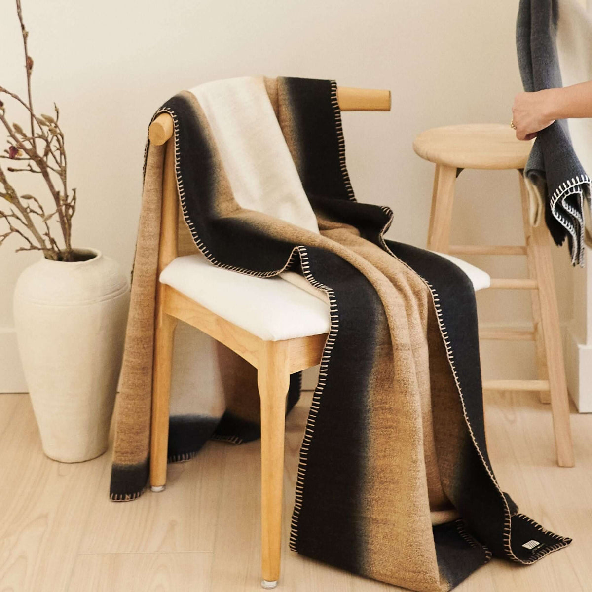 Siro Brown merino throw flowing over a pale upholstered chair beside a tall ceramic vase and wood stool