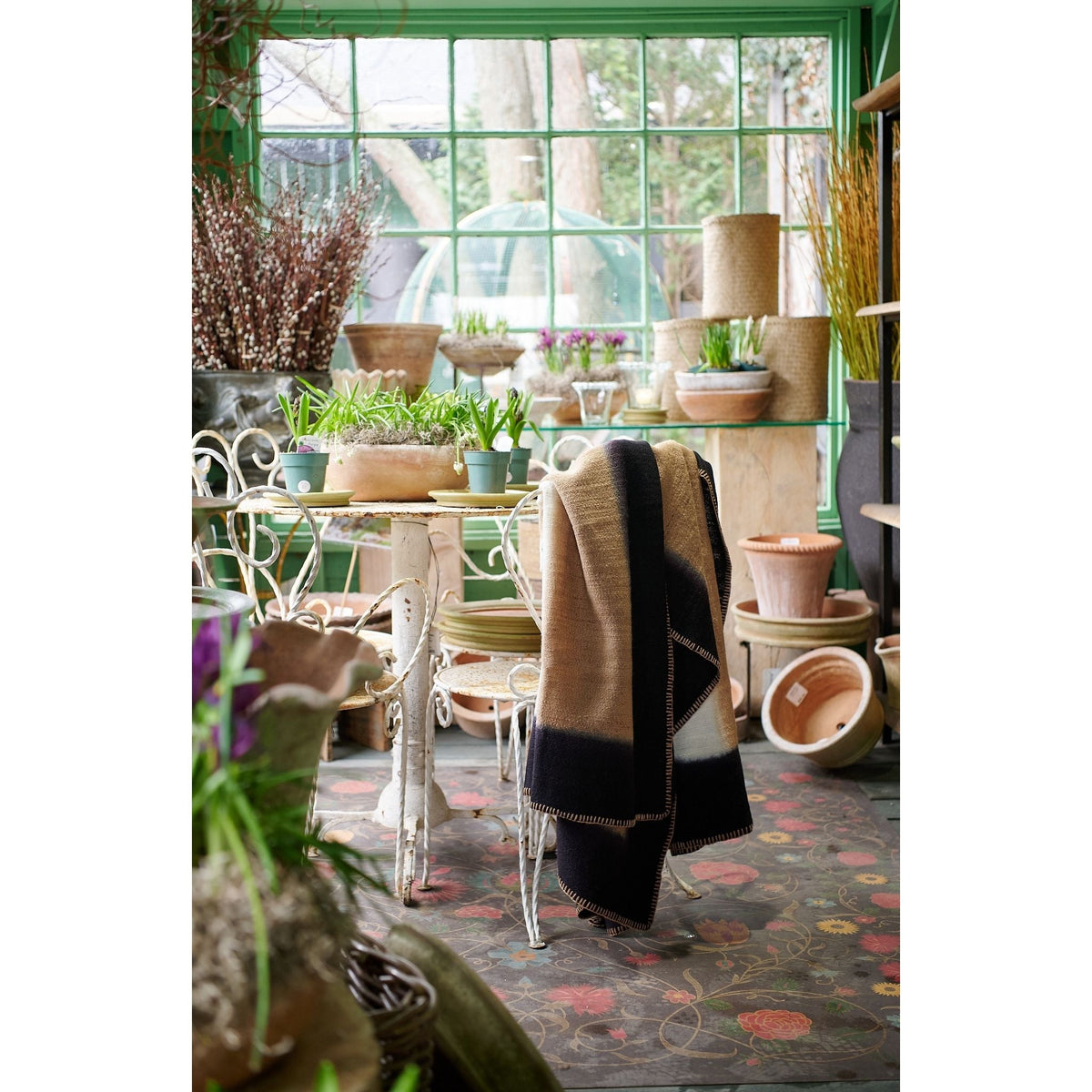 Siro Brown handwoven throw displayed over a chair among plants and pottery in a greenhouse-style shop