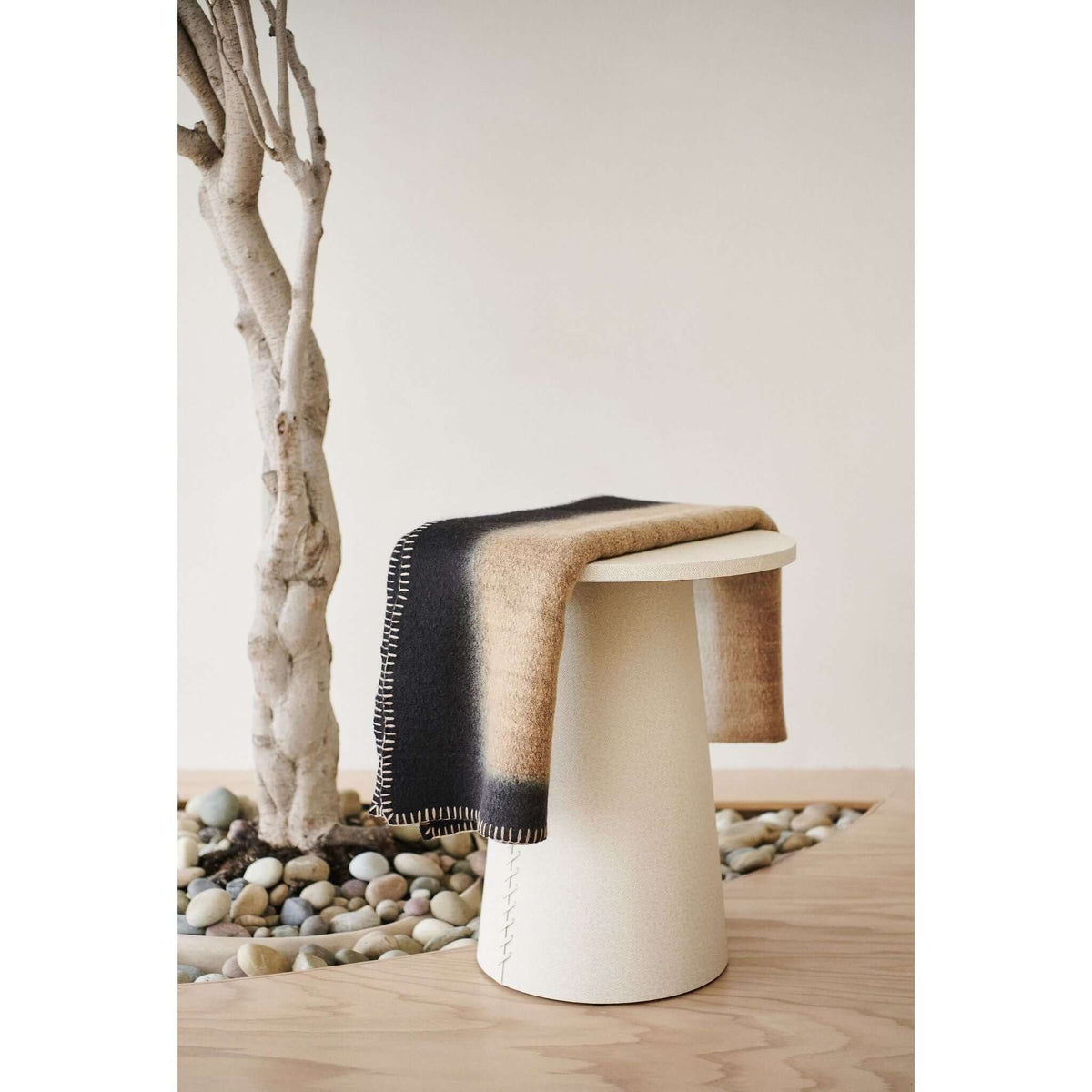 Siro Brown throw draped over a white stool beside a pale tree trunk and a bed of smooth decorative stones