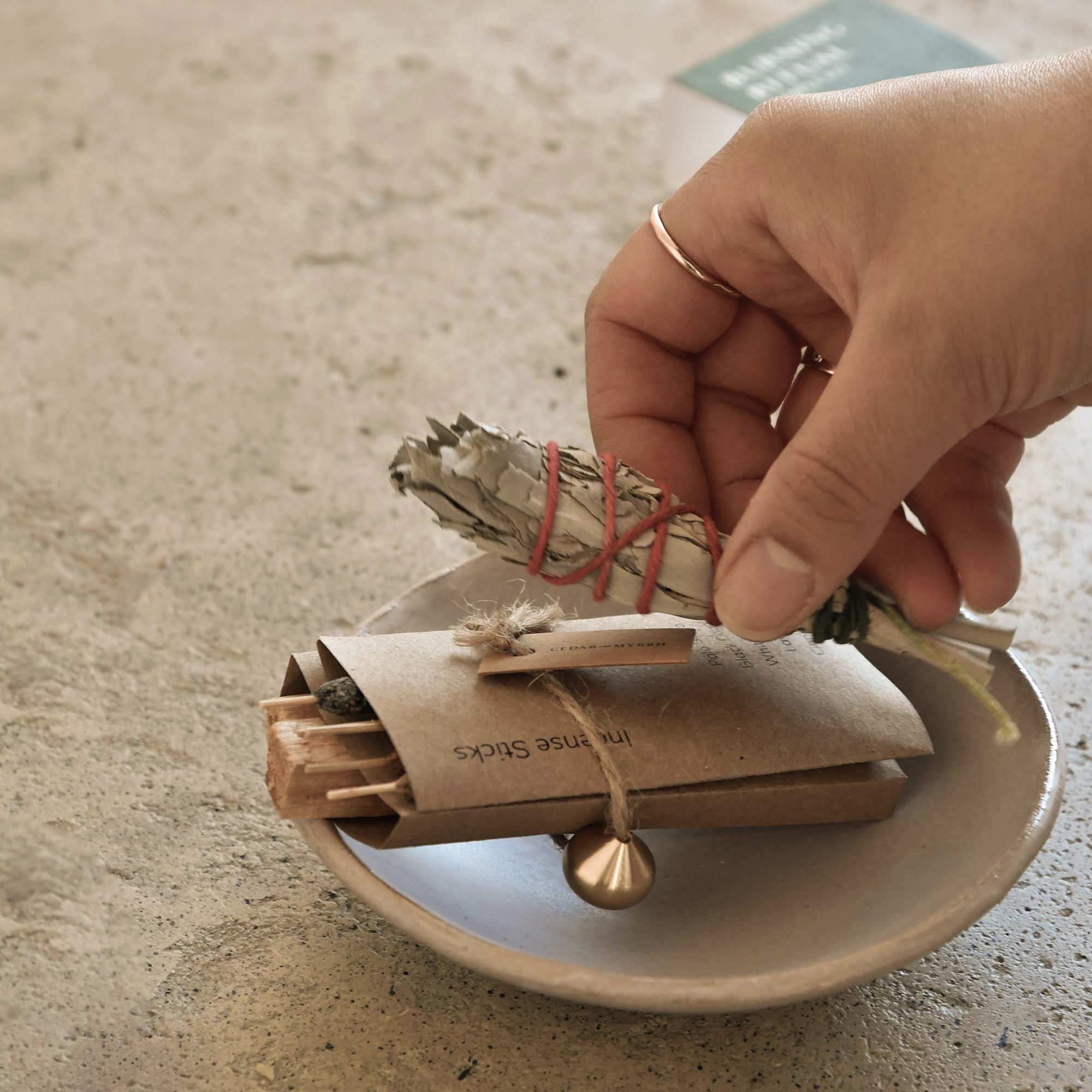 Hand holding sage smudge stick over a small dish with packaging on a neutral background