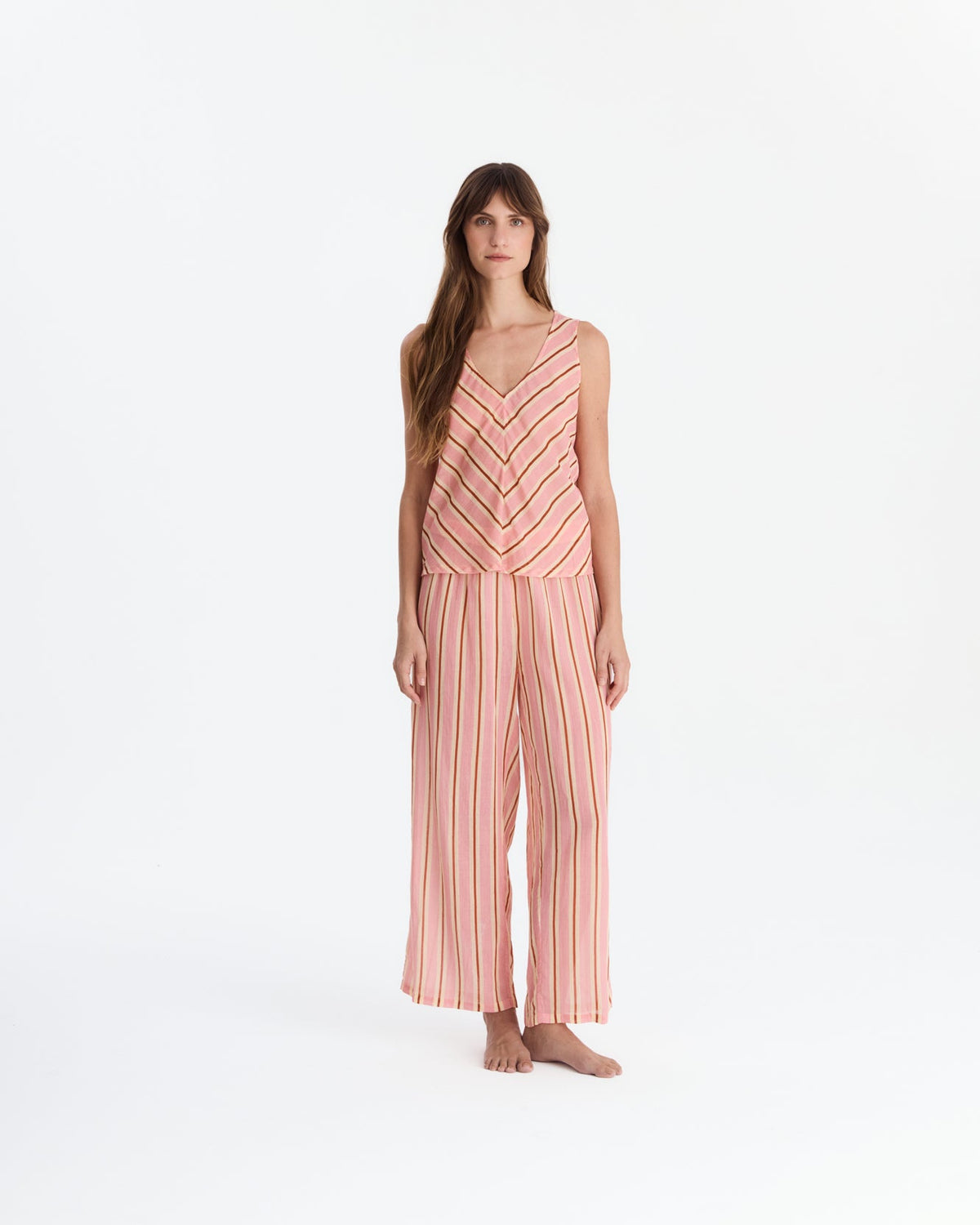 Barefoot model wearing coral pink pajamas with a sleeveless V-neck top featuring chevron stripes and matching vertically striped, wide-leg pants.