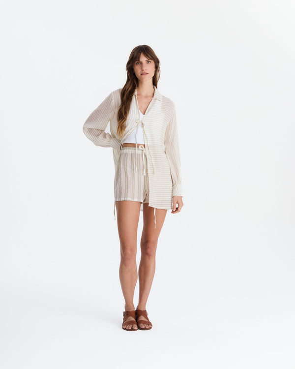 Model wearing a cream and beige striped cover-up with a collar, long sleeves, and front ties over a white crop top and striped shorts, paired with brown sandals.
