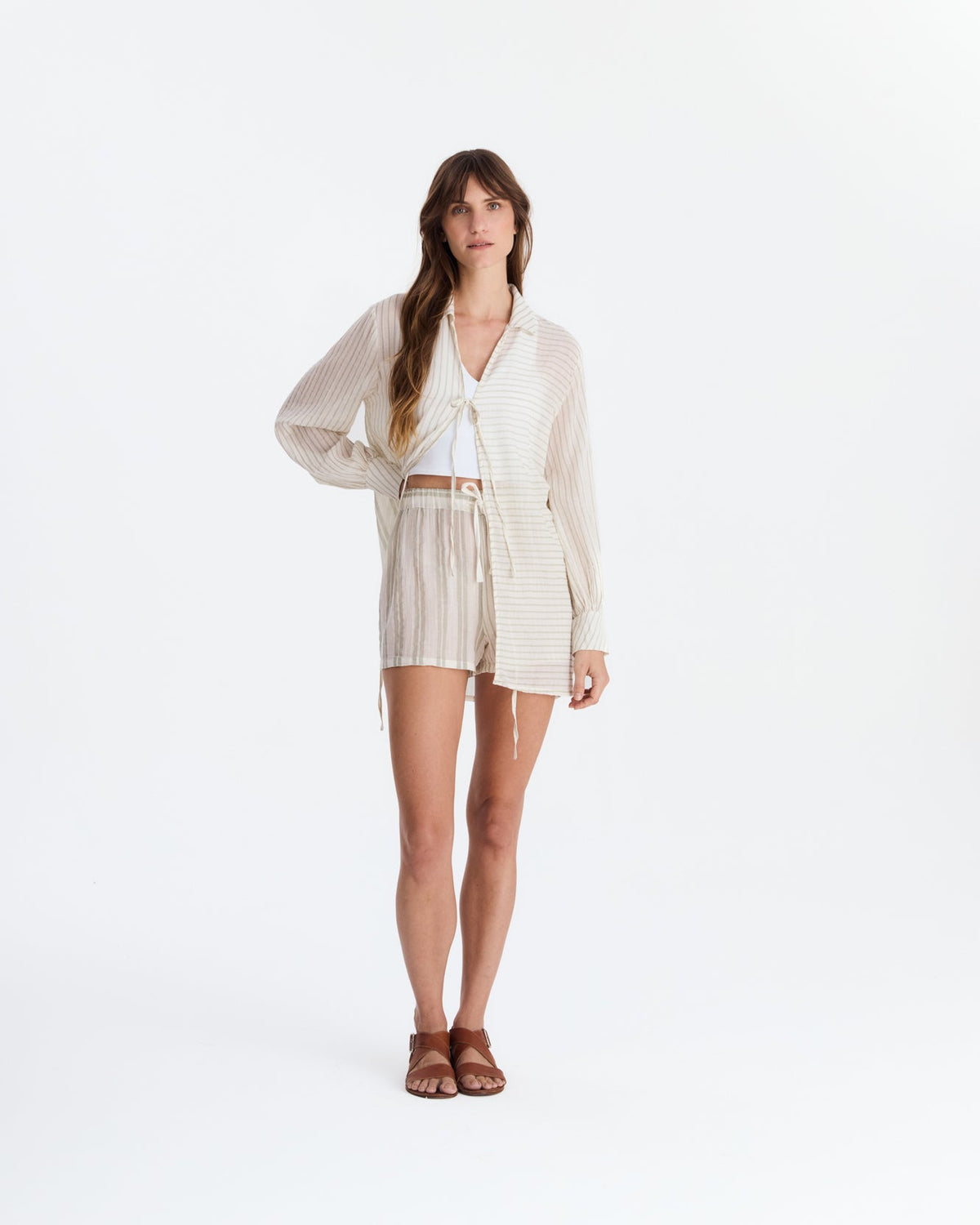Model wearing a cream and beige striped cover-up with a collar, long sleeves, and front ties over a white crop top and striped shorts, paired with brown sandals.