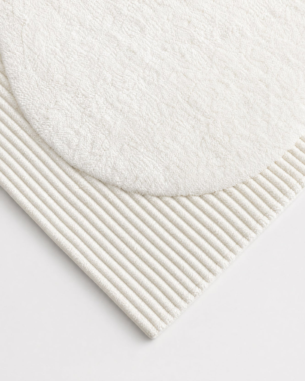 Close-up of the ivory bath rugβs corner, showing the curved edge of its plush oval center, parallel raised ribs, and neatly finished outer edge.