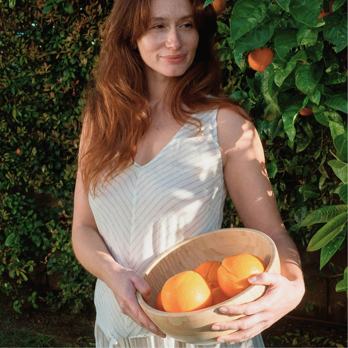 Closer outdoor view of the model holding a wooden bowl of oranges, showing the sleeveless top’s V-neck and diagonal cream and beige stripes.