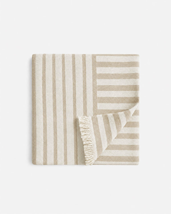Folded beige and cream Serena Beach Blanket with contrasting vertical and horizontal stripes, with one corner turned back to reveal cream fringe.
