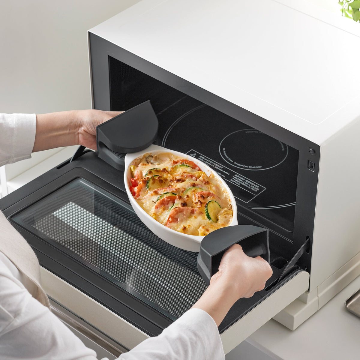 Person uses black Yamazaki Home Magnetic Pot Holders - Set of 2 - Silicone to remove an oval baking dish of browned food from a countertop oven.