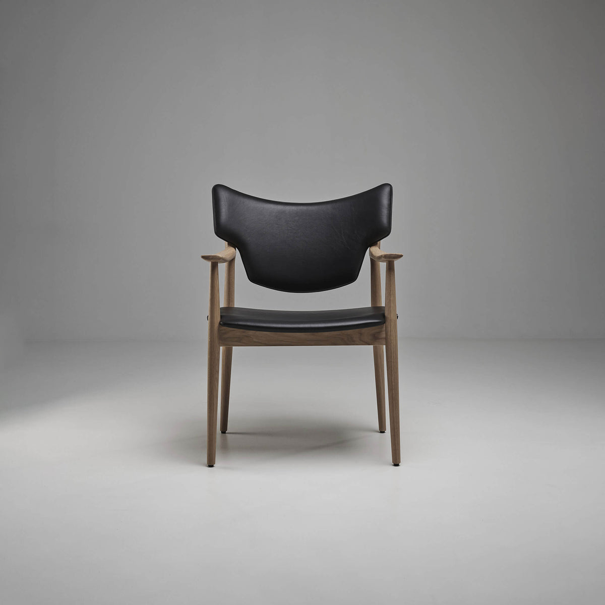 Front view of the black Eikund Veng Arm Chair in a gray studio, showing its curved upholstered back, separate seat, and slender brown wood frame.