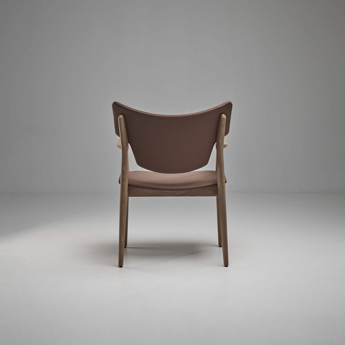 Rear view of the taupe-brown Eikund Veng Arm Chair in a gray studio, showing the smooth curved back panel, light brown wood uprights, and slim rear legs.