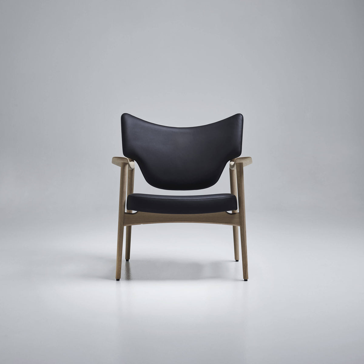 Front view of the Eikund Veng Lounge Chair with black upholstery and a light wood frame in a gray studio, showing the curved back, separate seat cushion, and tapered legs.