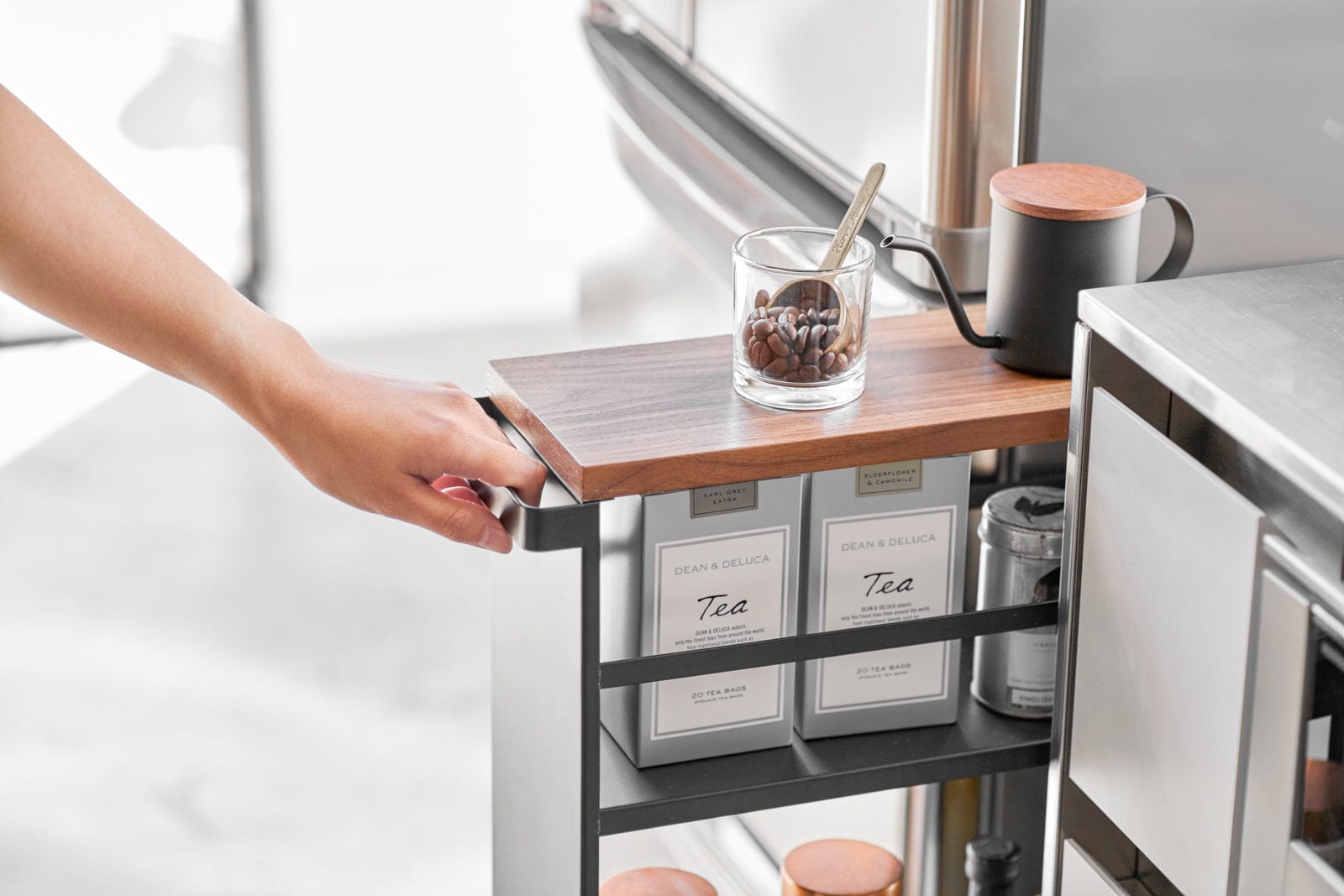 Kitchen cart with a wooden top holding a glass of coffee beans and a mug, next to tea bags.