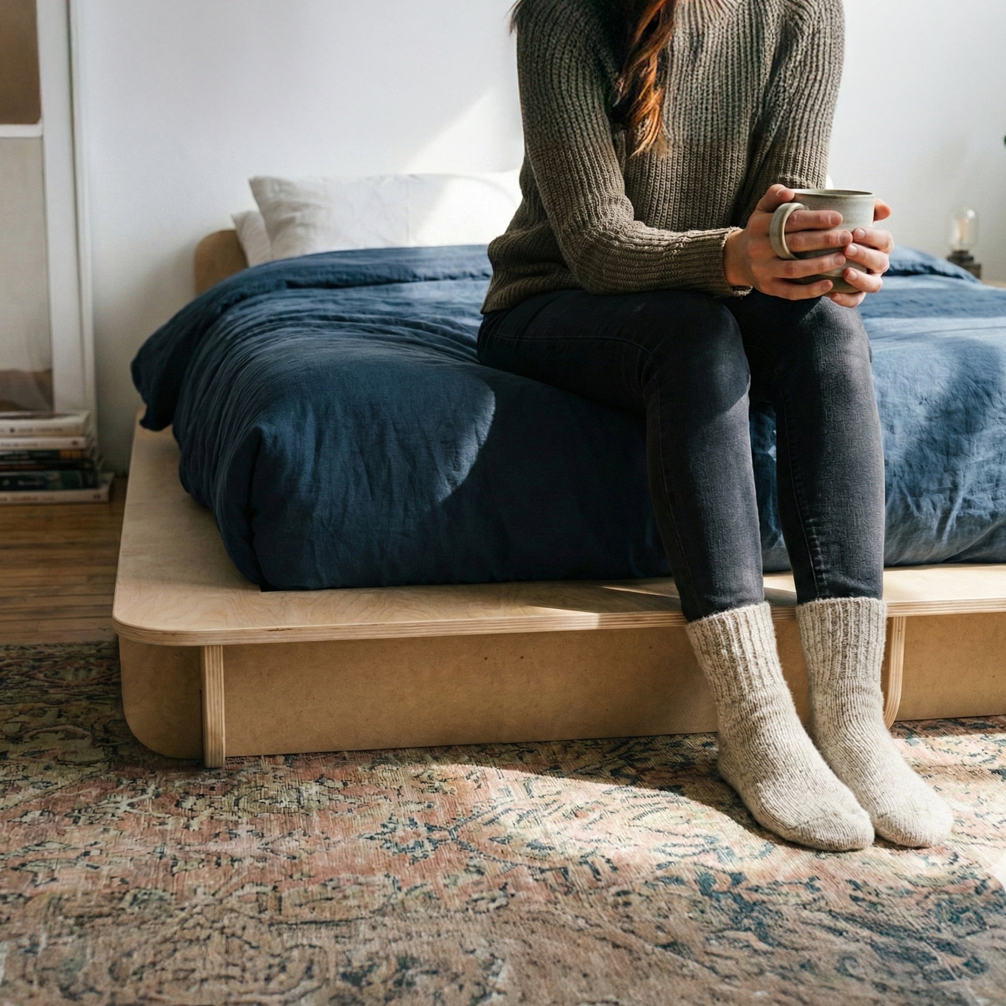 Person holding a mug while seated on the foot of a Studio Furnis bed platform with blue bedding