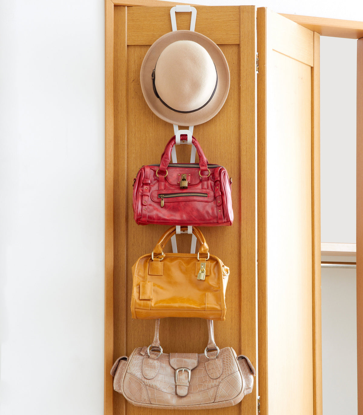 White Yamazaki Home Cascading Storage Hooks - Steel on a wooden slatted panel, displaying a beige hat above red, yellow and pale beige handbags.