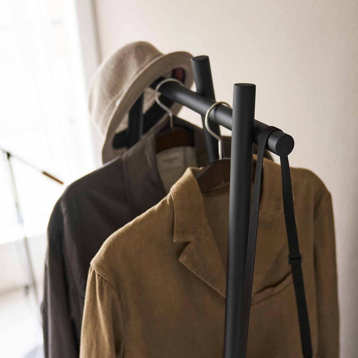 Close-up of the black Yamazaki Home Tri-Post Coat Rack (60" H) - Steel rail and upright posts, with wooden hangers, dark and brown coats, a neutral hat and a hanging bag strap.