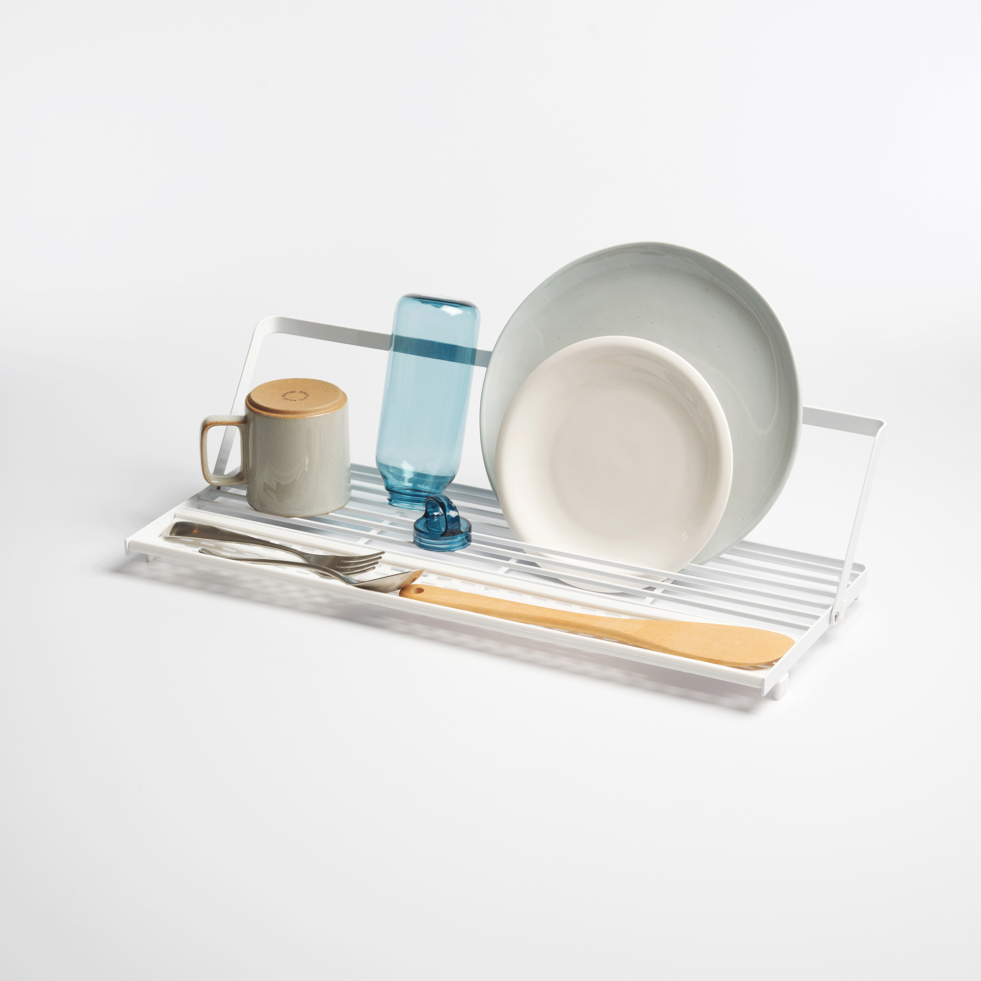 White Yamazaki Home Slim Over-the-Sink Dish Drying Rack - Steel lying flat with its support rail lowered, holding a mug, inverted bowls, cutlery and wooden utensils on a pale background.