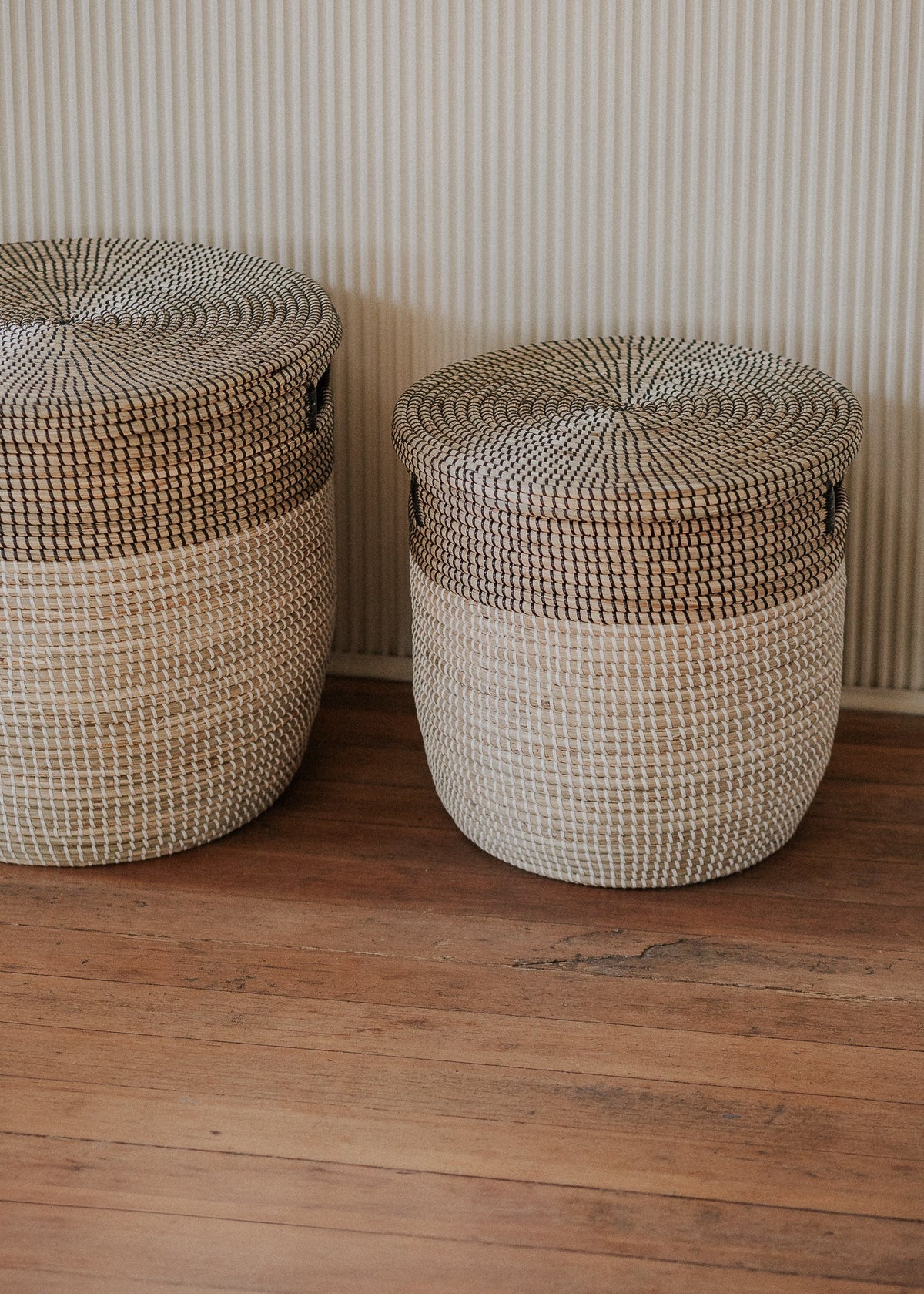 Pair of two-tone seagrass baskets arranged side by side against a softly striped wall.