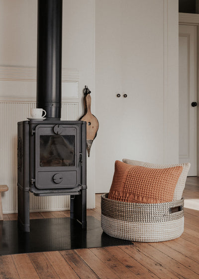 Black Medina floor basket holding a rust-colored cushion beside a black wood-burning stove.