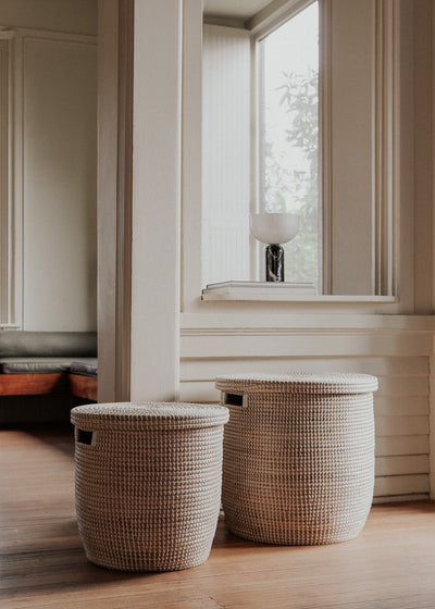 Two sizes of the lidded seagrass basket styled on a wood floor beside tall windows.