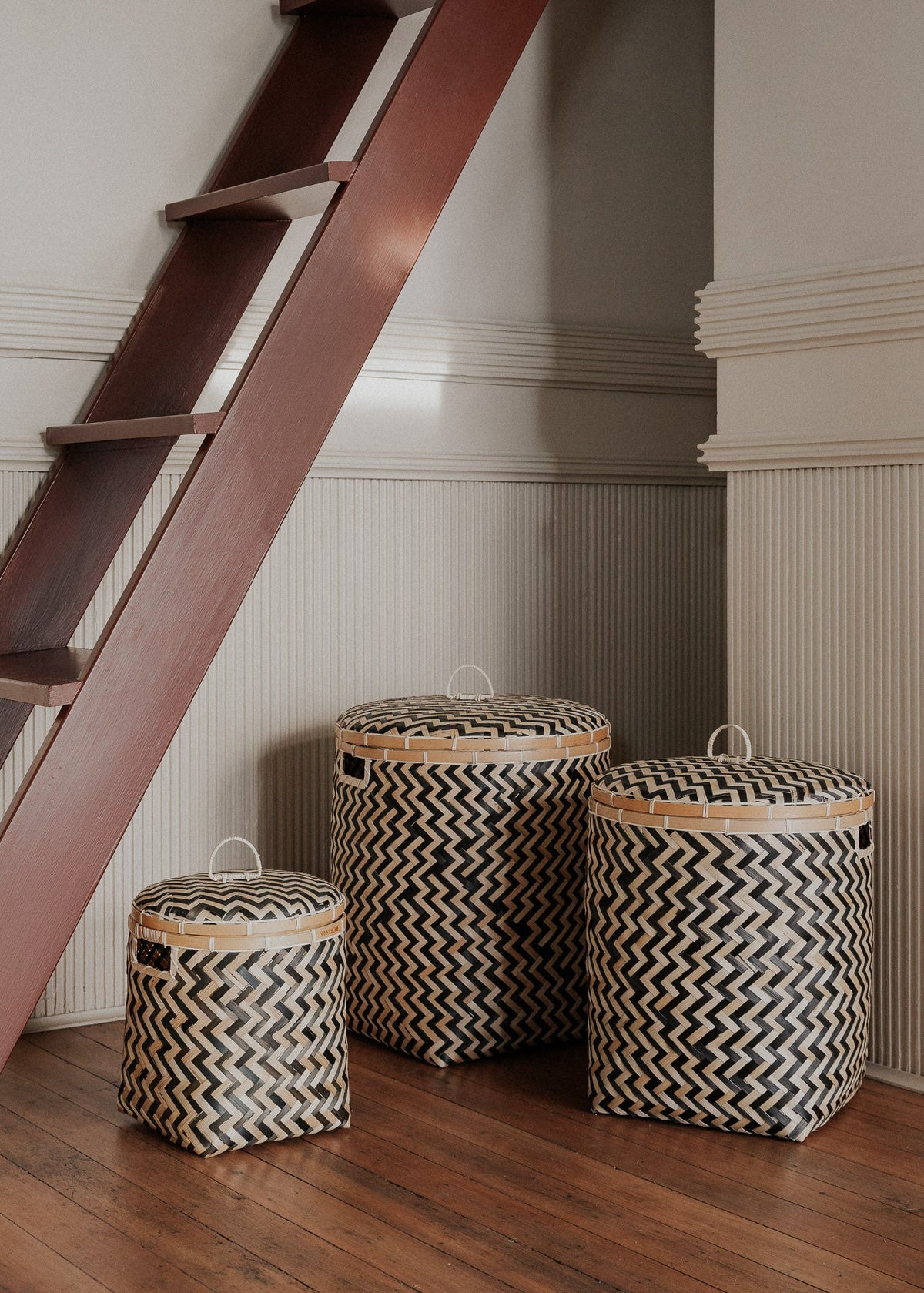 Three lidded Java baskets arranged beneath a wooden staircase on a dark wood floor.
