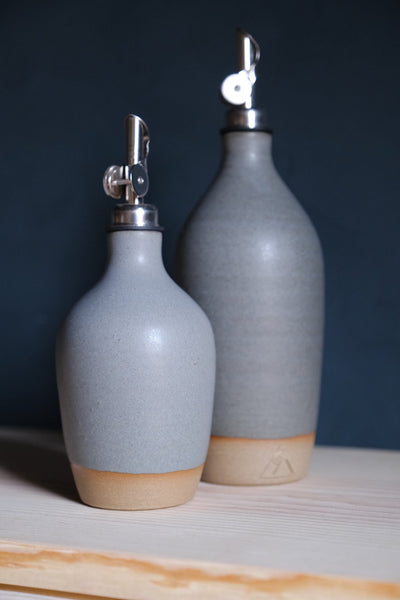 Light blue-grey Settle Ceramics Wander Oil Bottle in front of a taller slate-blue bottle on pale wood, showing fine speckles, horizontal ridges, metal pouring spouts and contrasting tan bases.