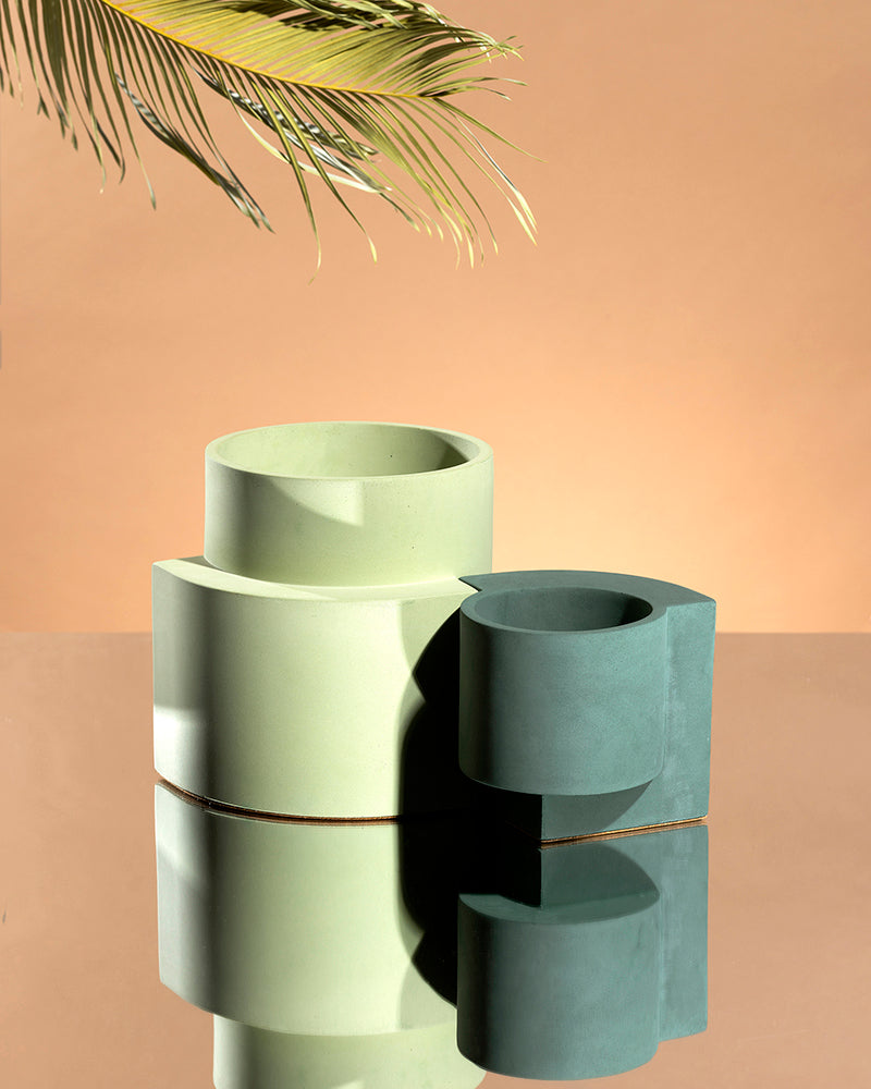 Dark blue-green Tortuga Forma Platform Planter Small - Polished Concrete beside a larger pale green planter on a mirrored surface, both empty, with reflections below and a leafy branch entering above against a peach background.