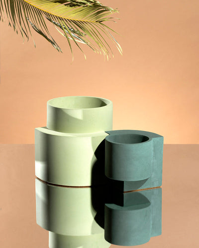 Dark blue-green Tortuga Forma Platform Planter Small - Polished Concrete beside a larger pale green planter on a mirrored surface, both empty, with reflections below and a leafy branch entering above against a peach background.