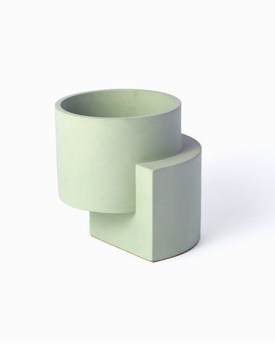 Pale green Tortuga Forma Platform Planter Medium - Polished Concrete viewed at an angle on white, showing an open cylindrical bowl raised above a curved vertical support, with a smooth surface and scattered tiny pores.