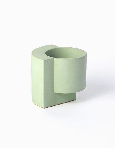 Pale green Tortuga Forma Platform Planter Small - Polished Concrete viewed at an angle on white, showing an empty cylindrical pot joined to a thick vertical slab extending along one side and beneath it, with tiny pores across the smooth surface.