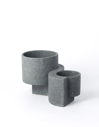 Graphite Tortuga Forma Platform Planter Set on white, with a larger pot behind a smaller one, showing pale terrazzo flecks, open cylindrical bowls and offset slab supports extending beneath them.