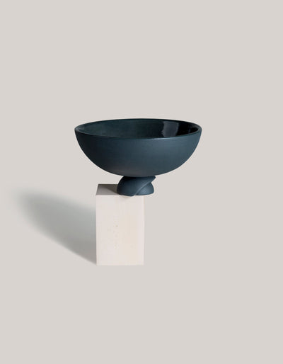 Graphite Tortuga Forma Kaolin Rice Bowl on a rectangular cream display block, viewed slightly from above to show its dark interior, rounded sides and overlapping curved foot, with a long shadow extending left.