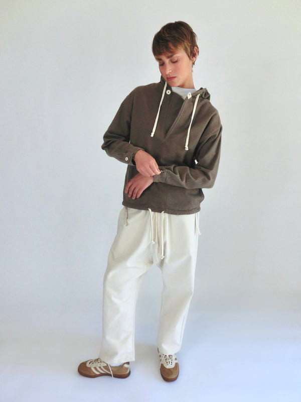 Model wearing a taupe-gray hooded Anorak with white drawcords, loose cream pants, and brown sneakers.