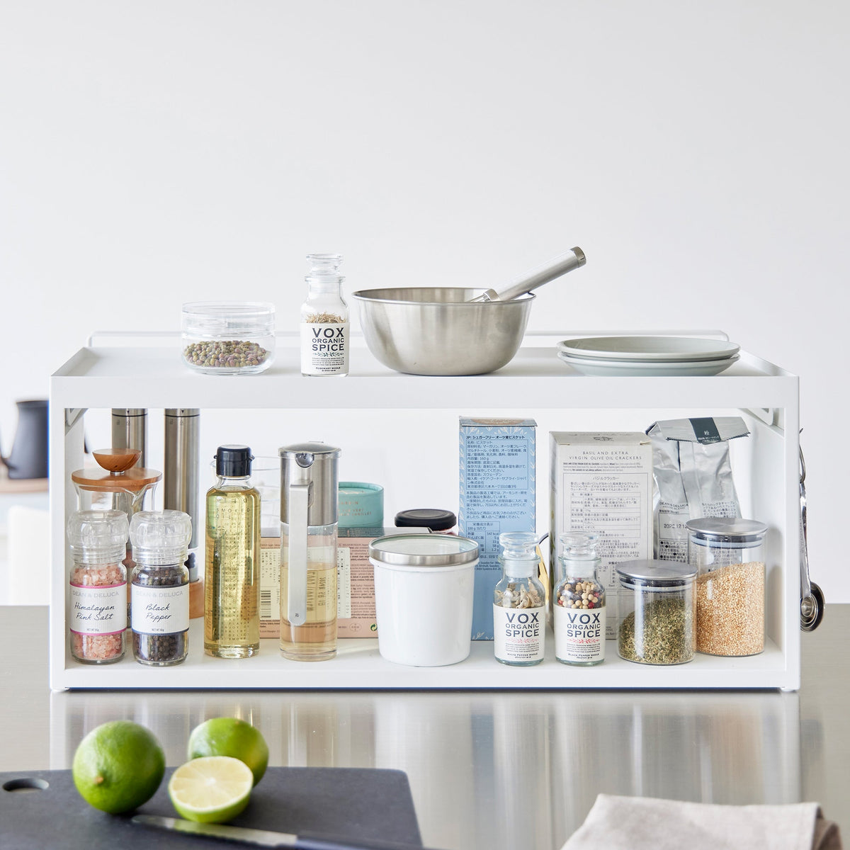 White Yamazaki Home Open-Back Countertop Storage Rack - Steel holding oils, seasonings, jars and canisters, with a saucepan and plates on top and cut limes on the counter in front.