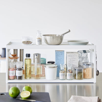 White Yamazaki Home Open-Back Countertop Storage Rack - Steel holding oils, seasonings, jars and canisters, with a saucepan and plates on top and cut limes on the counter in front.