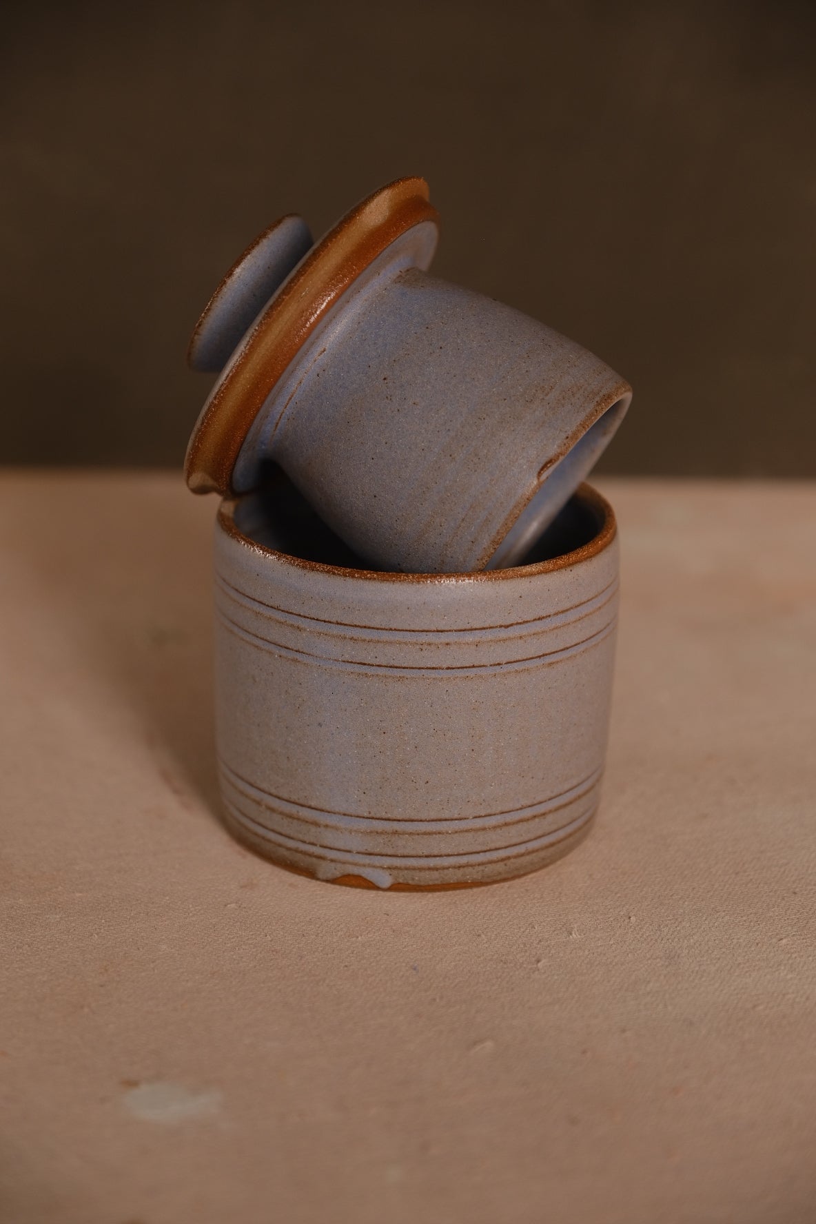Blue-grey Settle Ceramics Butterbell with pronounced horizontal bands and brown glaze accents, its cup-shaped lid resting diagonally across the open base.