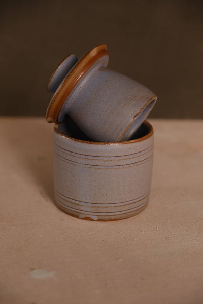 Blue-grey Settle Ceramics Butterbell with pronounced horizontal bands and brown glaze accents, its cup-shaped lid resting diagonally across the open base.