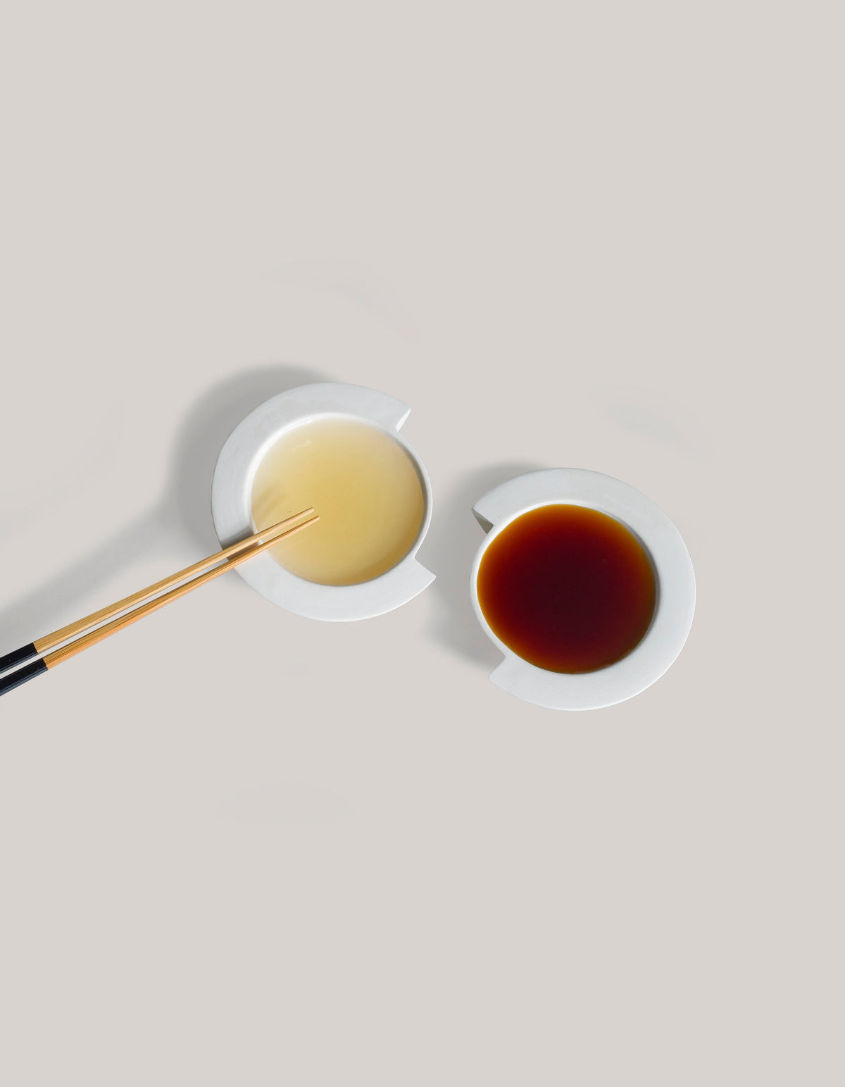 Overhead view of two white Tortuga Forma Kaolin Sauce Dishes holding pale yellow and dark brown liquids on a light grey surface, with chopsticks extending into one dish, showing shallow circular wells and broad rims interrupted by offset notches.