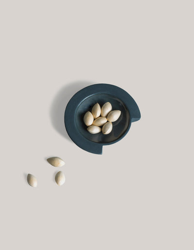 Graphite Tortuga Forma Kaolin Sauce Dish holding pale seeds, with several scattered beside it on a light grey surface, viewed overhead to show its round recessed centre and stepped rim.