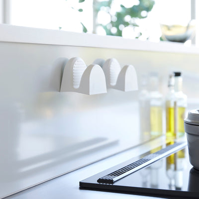 Two white Yamazaki Home Magnetic Pot Holders - Set of 2 - Silicone stored side by side on a kitchen backsplash, showing their arched profiles and ribbed inner surfaces.