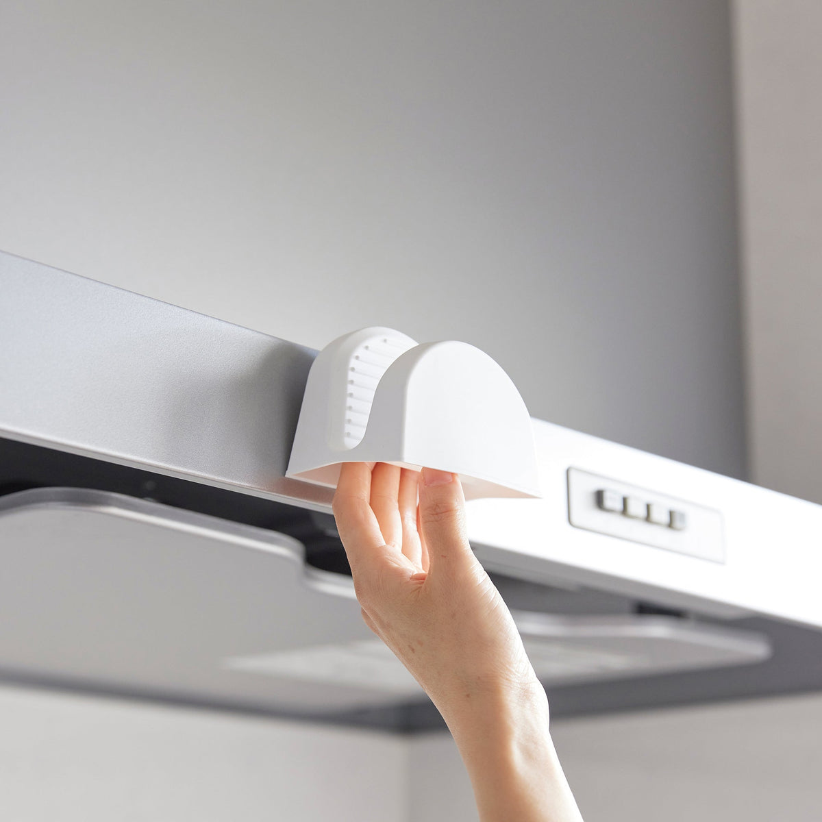 A hand reaches into a white Yamazaki Home Magnetic Pot Holder - Silicone attached to the front edge of a range hood, showing its open gripping pocket.